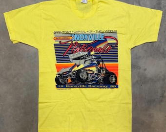 May include: Bright yellow t-shirt with a graphic of a sprint car, the text "Knoxville Nationals" and "19 Knoxville Raceway 89". The design incorporates blue, orange, and white. The shirt has a classic crew neck.