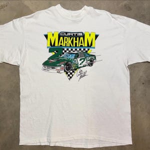 May include: White t-shirt featuring a graphic of a green race car with the number 2 and the name "Curtis Markham". The design includes the text "SKOL" and a signature. The shirt has short sleeves.