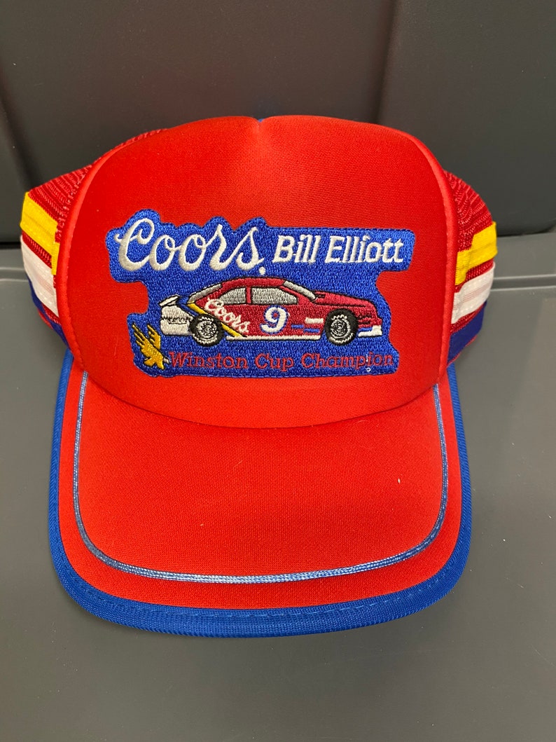 May include: Red, white, and blue trucker hat with a Coors Light logo and the text "Bill Elliott Winston Cup Champion". The hat features a small embroidered image of a race car with the number 9.