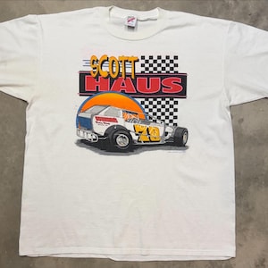 Puede incluir: Camiseta blanca con un gr&aacute;fico de coche de carreras y el texto "SCOTT HAUS" en rojo y amarillo. El dise&ntilde;o incluye una bandera a cuadros y un fondo de puesta de sol. El coche es blanco, azul y amarillo.