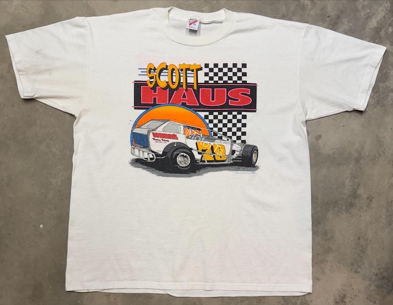 Puede incluir: Camiseta blanca con un gr&aacute;fico de coche de carreras y el texto "SCOTT HAUS" en rojo y amarillo. El dise&ntilde;o incluye una bandera a cuadros y un fondo de puesta de sol. El coche es blanco, azul y amarillo.