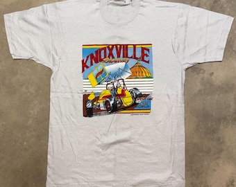 May include: A light gray short-sleeve t-shirt featuring a graphic print of a yellow and red sprint car on a dirt track. The text reads Knoxville Raceway and Sprint Car Capital of the World, with a yellow dome building in the background.