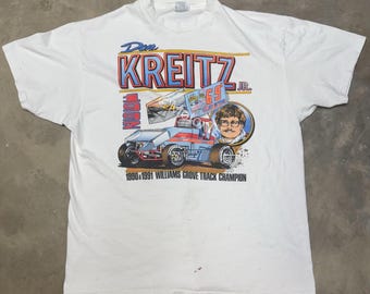 May include: White t-shirt featuring a colourful graphic of a race car driver named Don Kreitz Jr. The graphic includes a race car with the number 69 on it and the text "1990 & 1991 Williams Grove Track Champion".