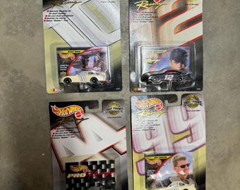 Op de afbeelding: Vier Hot Wheels NASCAR-modellauto's in hun originele verpakking. De auto's zijn wit met rode en blauwe accenten. De verpakking heeft het Hot Wheels-logo en de woorden "Test Track".