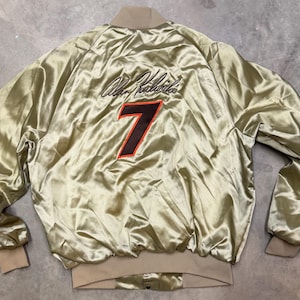 May include: A gold satin bomber jacket with a tan collar, cuffs, and hem. The back features the name "Alan Kulwicki" in black script above a large number "7" in orange with a black outline. The jacket has a shiny, wrinkled texture.