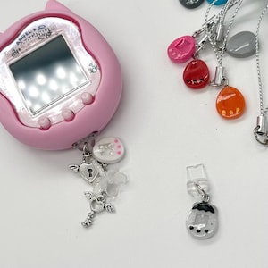 May include: A pink digital pet toy with a screen, buttons, and a charm. The charm includes a heart with a key, a white flower, and a small white circle with pink dots. Other charms are visible.