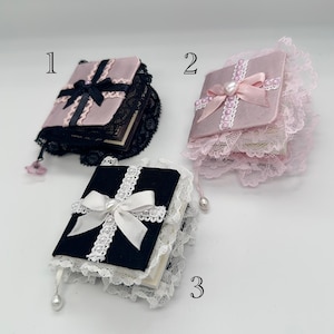 Puede incluir: Tres pequeños libros hechos a mano con ribetes de encaje y lazos de cinta. Los libros son de color rosa, negro y blanco. Los libros están numerados del 1 al 3.