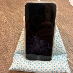 May include: A smartphone in a clear protective case resting on a wedge-shaped fabric stand. The stand features a light blue and white diamond pattern with small red dots at the intersections and includes a light blue fabric loop at the top.