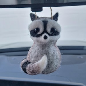 Könnte beinhalten: Ein Filz-Waschbär-Ornament mit schwarzer Maske, schwarzen Ohren und einem gestreiften Schwanz. Der Waschbär hat ein weißes Gesicht und eine weiße Brust und hängt an einer Schnur. Das Ornament hängt am Rückspiegel eines Autos.