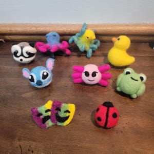 Può includere: Una collezione di figure di animali in feltro fatte a mano, tra cui un procione, un polpo, un'anatra, una rana, una coccinella e un personaggio simile a Stitch. Le figure sono in vari colori, come blu, rosa, giallo e verde.