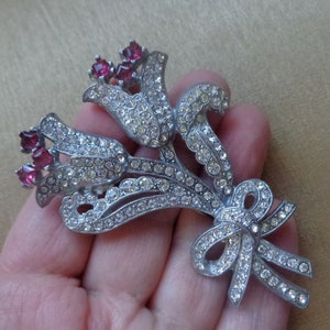 Può includere: Un broche color argento con un bouquet di tre tulipani stilizzati con strass rosa. I tulipani sono legati insieme da un fiocco.