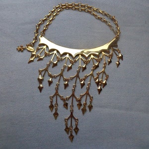 Puede incluir: Un collar dorado con un diseño único. El collar presenta una sección superior curva con una serie de ramas estilizadas colgantes, cada una adornada con colgantes en forma de diamante. La cadena es una doble hilera de pequeños círculos enlazados.