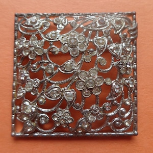May include: A silver square brooch with a delicate floral design and clear rhinestones.