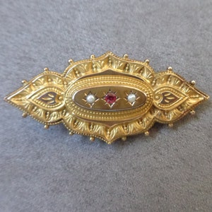 May include: A gold brooch with an oval centre featuring a red gemstone surrounded by two white pearls. The brooch has a detailed, ornate design.