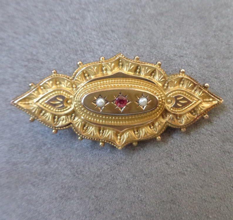 May include: A gold brooch with an oval centre featuring a red gemstone surrounded by two white pearls. The brooch has a detailed, ornate design.