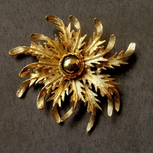 May include: A gold-toned brooch in the shape of a stylised flower with a round, raised centre. The flower has many petals that are textured and have a slightly jagged edge.