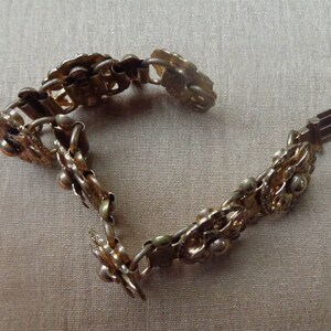 May include: A vintage gold-toned bracelet with a floral design. The bracelet is composed of interconnected floral and leaf-shaped links, with small spherical accents. The clasp is visible at the end of the bracelet. The bracelet is displayed on a neutral-colored surface.