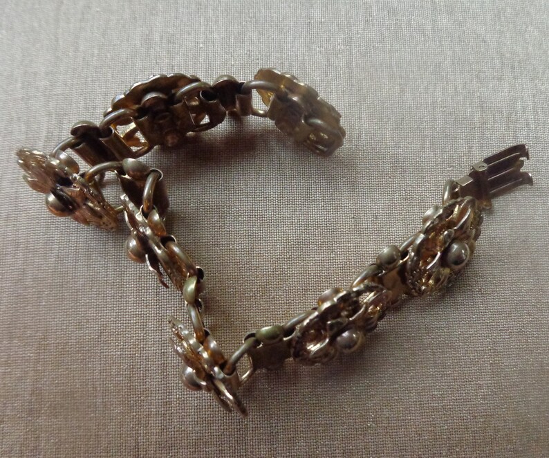 May include: A vintage gold-toned bracelet with a floral design. The bracelet is composed of interconnected floral and leaf-shaped links, with small spherical accents. The clasp is visible at the end of the bracelet. The bracelet is displayed on a neutral-colored surface.