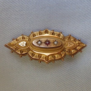 May include: A gold brooch with an oval-shaped centre featuring a red stone surrounded by two white pearls. The brooch has a detailed, ornate design with a scalloped edge.