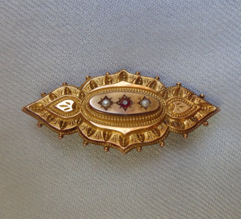 May include: A gold brooch with an oval-shaped centre featuring a red stone surrounded by two white pearls. The brooch has a detailed, ornate design with a scalloped edge.