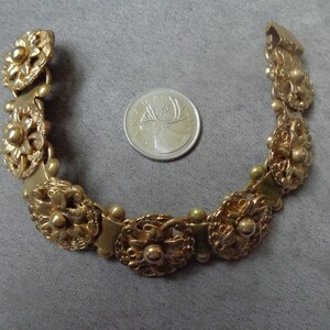 May include: A gold-toned bracelet with floral and beaded details. The bracelet is composed of several ornate, flower-shaped links connected by smaller links. A Canadian quarter coin is next to the bracelet for size comparison.