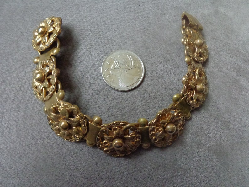 May include: A gold-toned bracelet with floral and beaded details. The bracelet is composed of several ornate, flower-shaped links connected by smaller links. A Canadian quarter coin is next to the bracelet for size comparison.