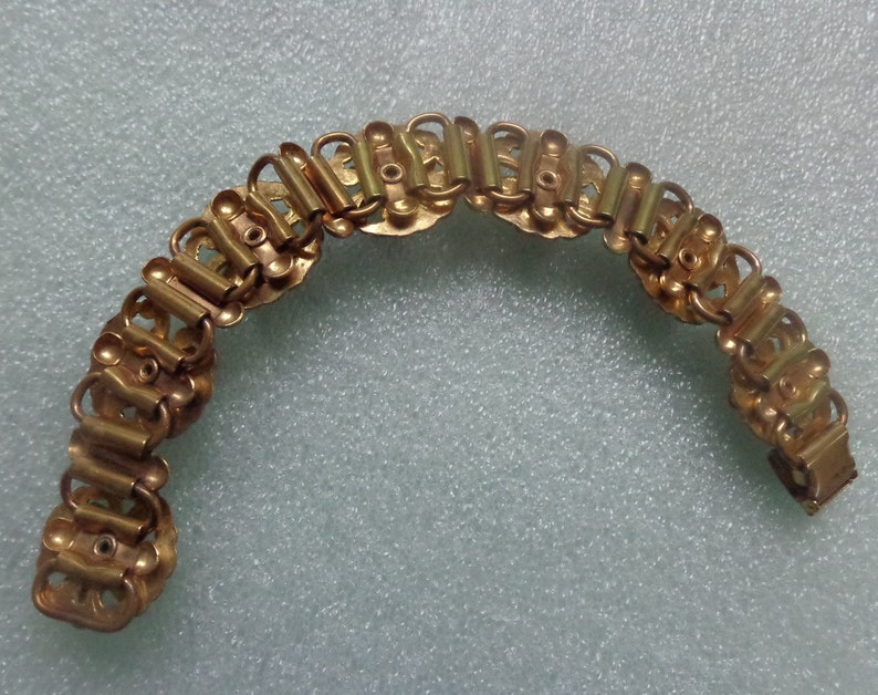 May include: A gold-toned bracelet with a repeating pattern of linked rectangular and circular elements. The bracelet is curved, suggesting it is flexible and designed to fit the wrist. The metal has a polished finish.