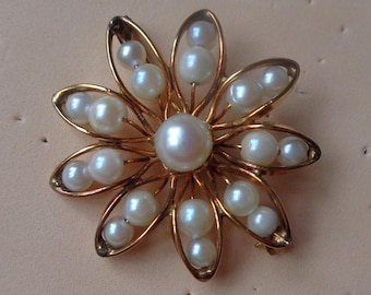 May include: A gold-toned flower brooch featuring a central large pearl surrounded by smaller pearls within the petals. The brooch has a delicate, openwork design, creating a floral pattern. The pearls are white and have a lustrous sheen.