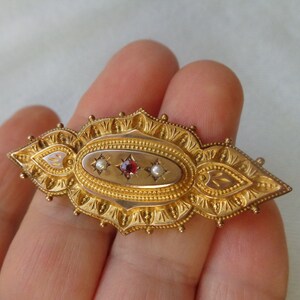 May include: A gold brooch with an oval centre featuring a red gemstone and two white pearls. The brooch is decorated with intricate floral designs.