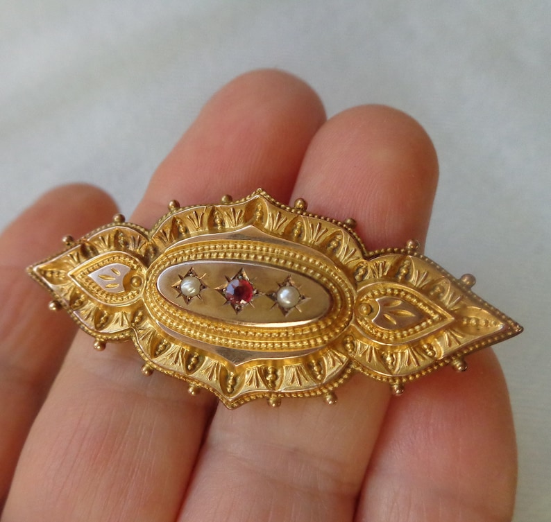 May include: A gold brooch with an oval centre featuring a red gemstone and two white pearls. The brooch is decorated with intricate floral designs.