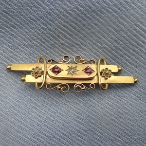 Puede incluir: Un broche dorado con una pieza central rectangular engastada con dos piedras rojas y una sola piedra blanca. El broche está decorado con intrincados trabajos de volutas.