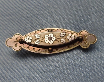 May include: A silver and gold brooch with a floral design. The brooch is oval shaped and has a white flower with leaves in the center. The brooch is surrounded by a decorative border.