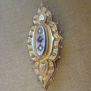 May include: A gold brooch with an oval centre featuring three red gemstones and two white pearls. The brooch is decorated with intricate floral designs.