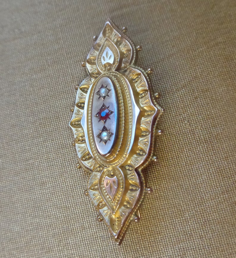 May include: A gold brooch with an oval centre featuring three red gemstones and two white pearls. The brooch is decorated with intricate floral designs.
