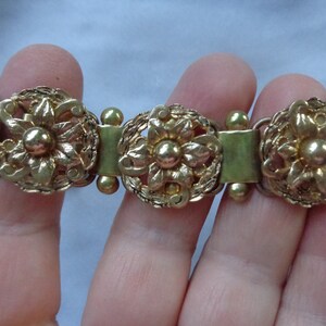 May include: Gold-toned bracelet featuring three ornate floral medallions. Each medallion has a central flower surrounded by detailed filigree and connected by rectangular and spherical links. The bracelet is a vintage style.