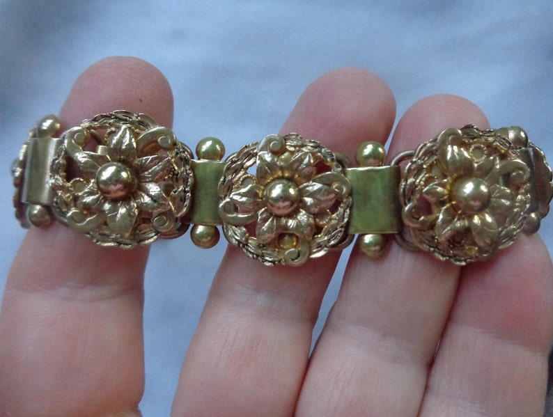 May include: Gold-toned bracelet featuring three ornate floral medallions. Each medallion has a central flower surrounded by detailed filigree and connected by rectangular and spherical links. The bracelet is a vintage style.