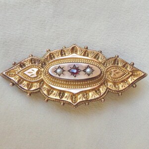 May include: A gold brooch with an oval centre featuring three red gemstones and two pearls. The brooch is decorated with intricate floral designs.