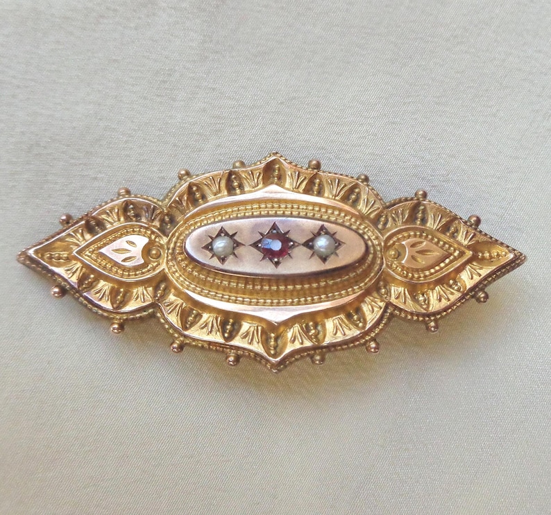 May include: A gold brooch with an oval centre featuring three red gemstones and two pearls. The brooch is decorated with intricate floral designs.