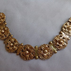 May include: Gold-toned bracelet featuring ornate floral designs. The bracelet is composed of multiple circular, flower-shaped links connected by smaller, rounded beads and rectangular segments. The clasp is a simple, rectangular design.