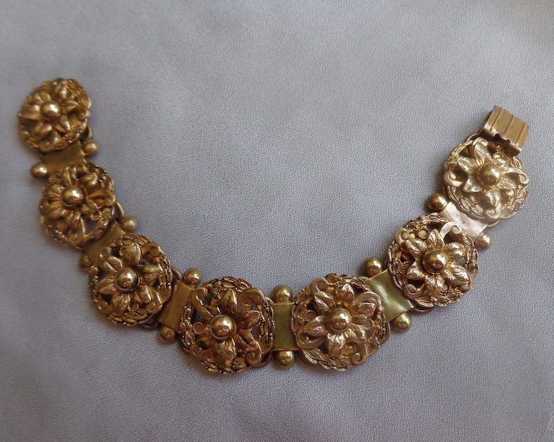 May include: Gold-toned bracelet featuring ornate floral designs. The bracelet is composed of multiple circular, flower-shaped links connected by smaller, rounded beads and rectangular segments. The clasp is a simple, rectangular design.