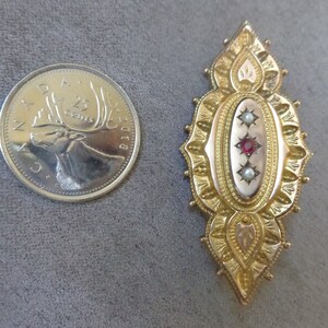 May include: A gold-toned brooch with an oval centre featuring a red stone and two white pearls. The brooch is intricately detailed with a floral design.