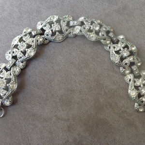 Puede incluir: Un brazalete plateado con un diseño delicado e intrincado adornado con brillantes transparentes. El brazalete está hecho de metal y tiene un cierre de broche.