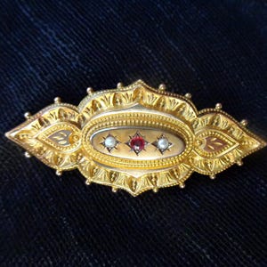 May include: A gold brooch with an oval-shaped centre featuring three gemstones: two white pearls and a red ruby. The brooch is intricately designed with a detailed floral pattern.