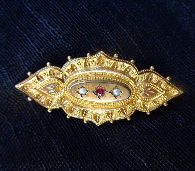 May include: A gold brooch with an oval-shaped centre featuring three gemstones: two white pearls and a red ruby. The brooch is intricately designed with a detailed floral pattern.