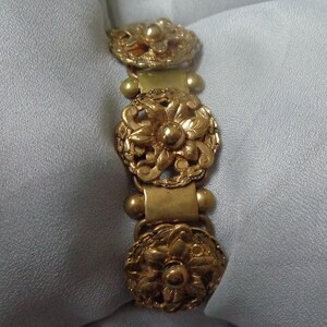 May include: Gold-toned bracelet featuring three ornate floral medallions. The bracelet has a series of rectangular links and small spherical accents. The floral designs are detailed with intricate petal and leaf patterns, creating a vintage aesthetic.