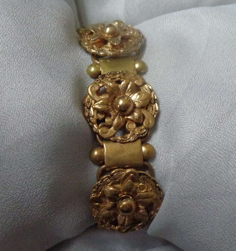 May include: Gold-toned bracelet featuring three ornate floral medallions. The bracelet has a series of rectangular links and small spherical accents. The floral designs are detailed with intricate petal and leaf patterns, creating a vintage aesthetic.