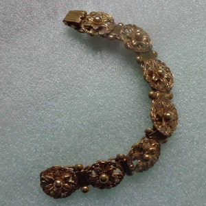 May include: A gold-toned bracelet featuring a series of ornate, flower-shaped links. The bracelet has a delicate, filigree design with small, round accents between the floral elements. The clasp is a simple, rectangular design.