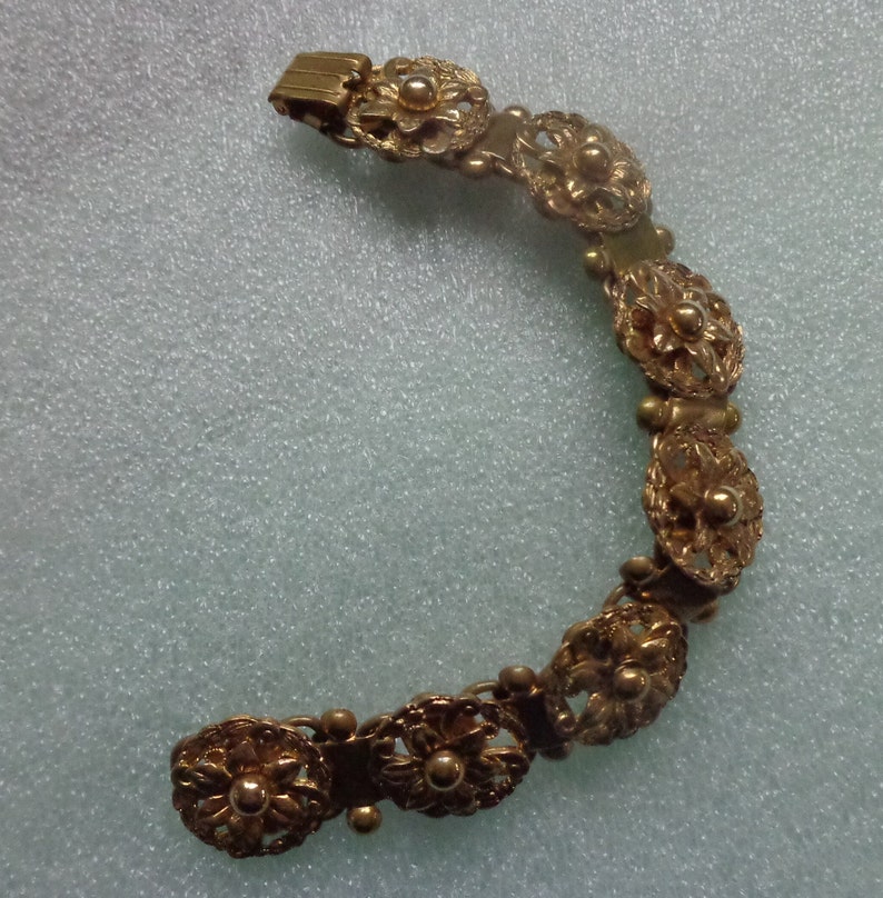 May include: A gold-toned bracelet featuring a series of ornate, flower-shaped links. The bracelet has a delicate, filigree design with small, round accents between the floral elements. The clasp is a simple, rectangular design.