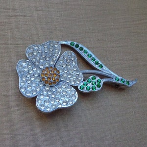 May include: A silver flower brooch with clear and amber rhinestones. The flower has a green stem with green rhinestones.