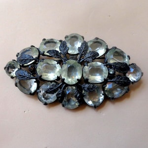 May include: An oval brooch with light green, oval and rectangular gemstones set in a dark metal frame. The design includes leaf accents. The brooch features a small, round gemstone. The brooch is a vintage style.